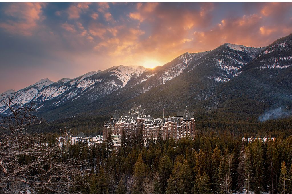 A picture of Fairmont Springs Banff Hotel, which is one of the two most well-known hotels in Banff. The hotel lies along Bow River and is surrounded by evergreen trees and snowy mountains.