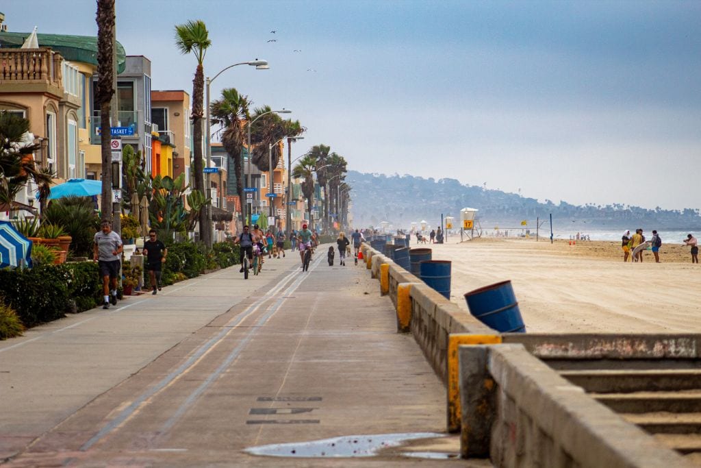 A picture of the pathway at Mission Beach. People can be seen jogging, walking, and riding along the pathway. There are also lots of colorful houses and lifeguard towers flanking the pathway.