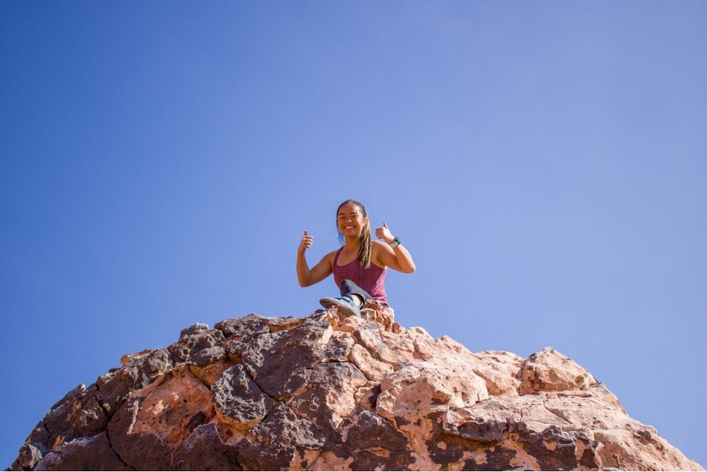 A picture of Kristin giving two thumbs up on top of a boulder she climbed. Climbing is one of the many reasons people find Red Rock Canyon worth visiting.