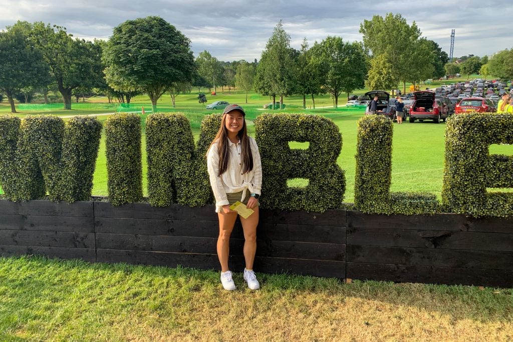 A picture of Kristin smiling as she stands in front of the grass display that spells Wimbledon.
