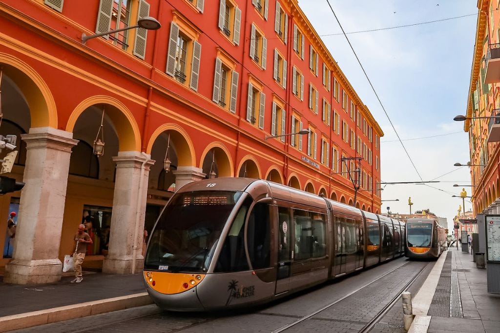 A picture of the trams passing through the bright salmon colored buildings near Place Massena in Nice France.