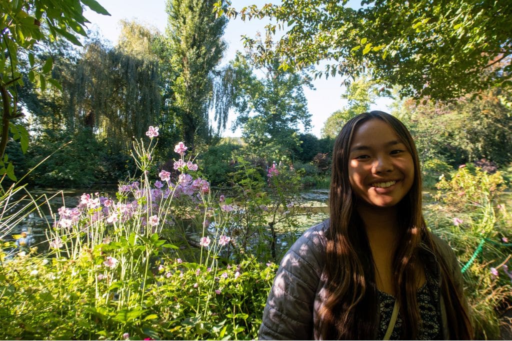 A picture of Kristin smiling while standing underneath a tree next to the water lily pond in Monet's Gardens. Definitely take a few moments to admire the beautiful water lilies if you do a Day Trip from Paris to Giverny.