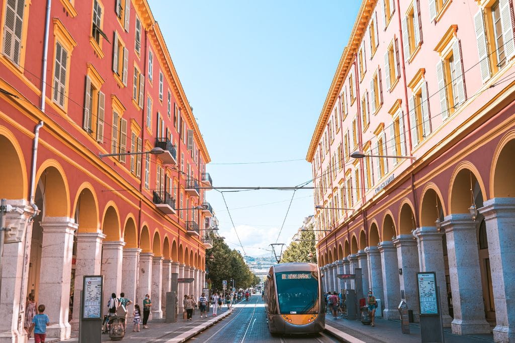 A picture of the tram in Nice as it moves between two pink buildings. The public transport is amazing and a great way to ensure you see everything you want to during your one day in nice France.