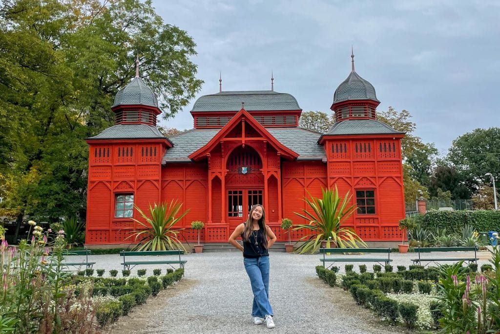 A picture of Kristin posing in front of a bright red building that can be found at the Zagreb Botanical Gardens. If you spend 3 Days in Zagreb Croatia during the summer, make sure to walk through these lovely gardens.