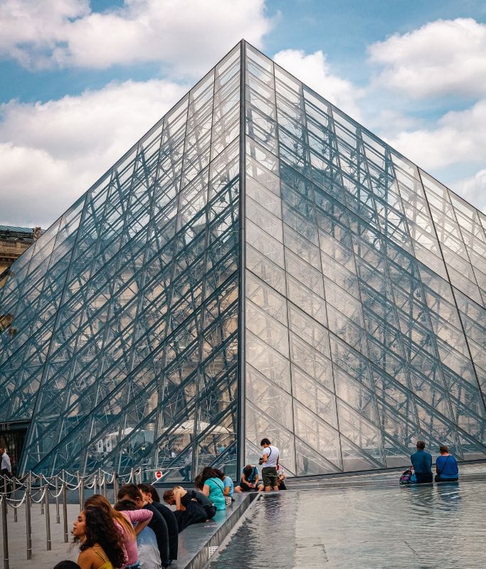 A picture of people sitting on fountain ledge outside of the Louvre. You can see the iconic glass pyramid.