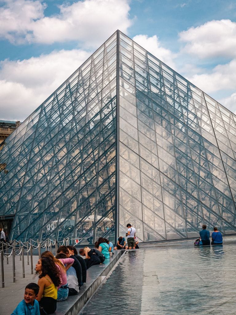 A picture of people sitting on fountain ledge outside of the Louvre. You can see the iconic glass pyramid.