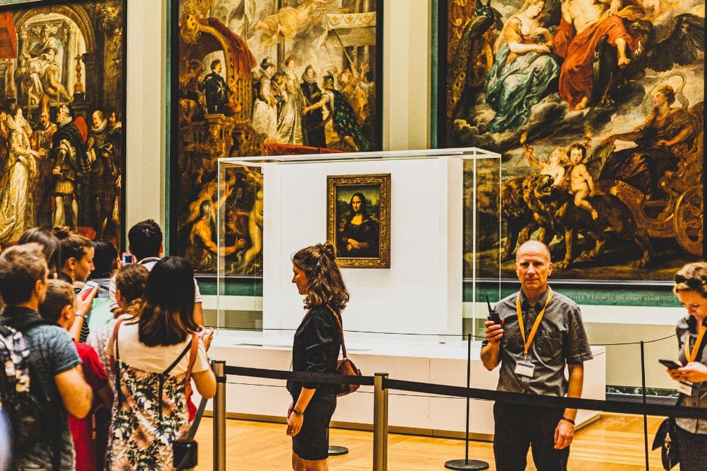 A picture of the Mona Lisa inside the louvre. You can see a very serious man and lots of people. To avoid Paris Syndrome, expect some French people to not be so friendly.