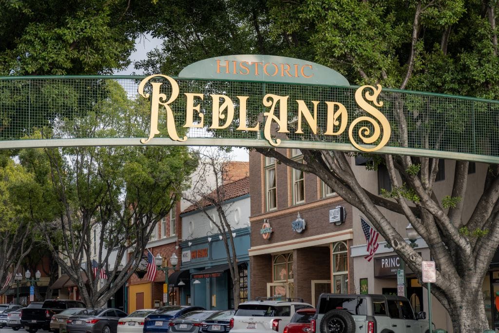 A picture of the sign that marks the entrance to downtown Redlands. In the background or several storefronts. 