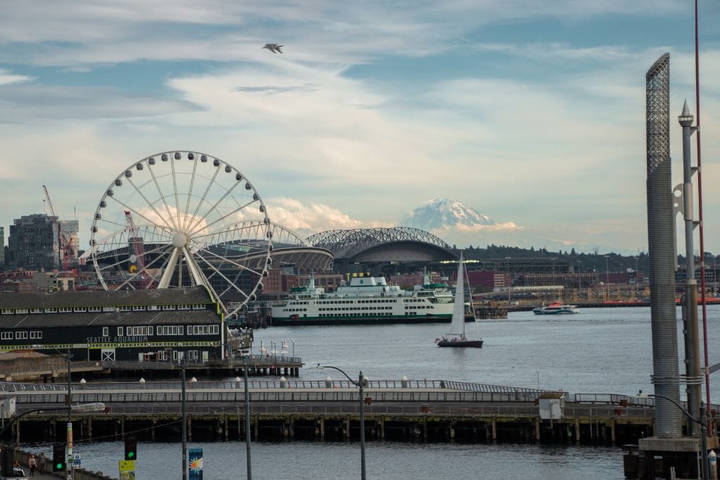 A picture of the Seattle harbor with the giant ferris wheel and surrounding mountains in the picture as well