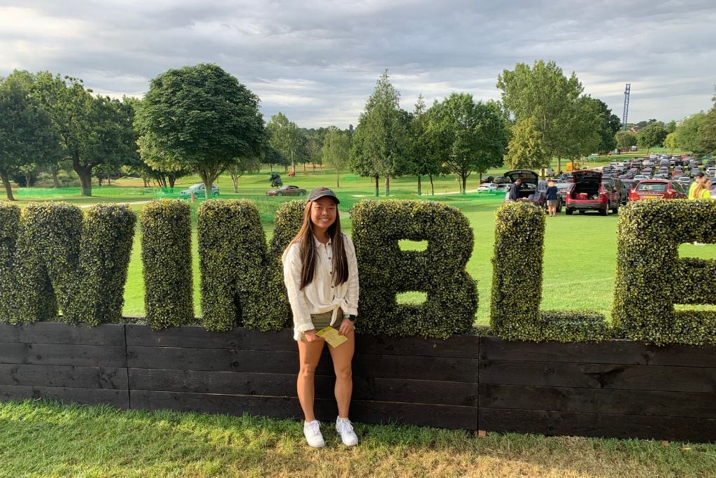 A picture of Kristin smiling in front of the Wimbledon grass sign.