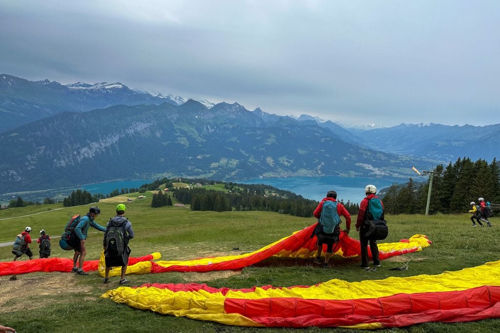 A picture taken from the launch site in Beatenberg. In this picture, there are lots of guests and their pilots getting ready for take off with their red and yellow paragliders.