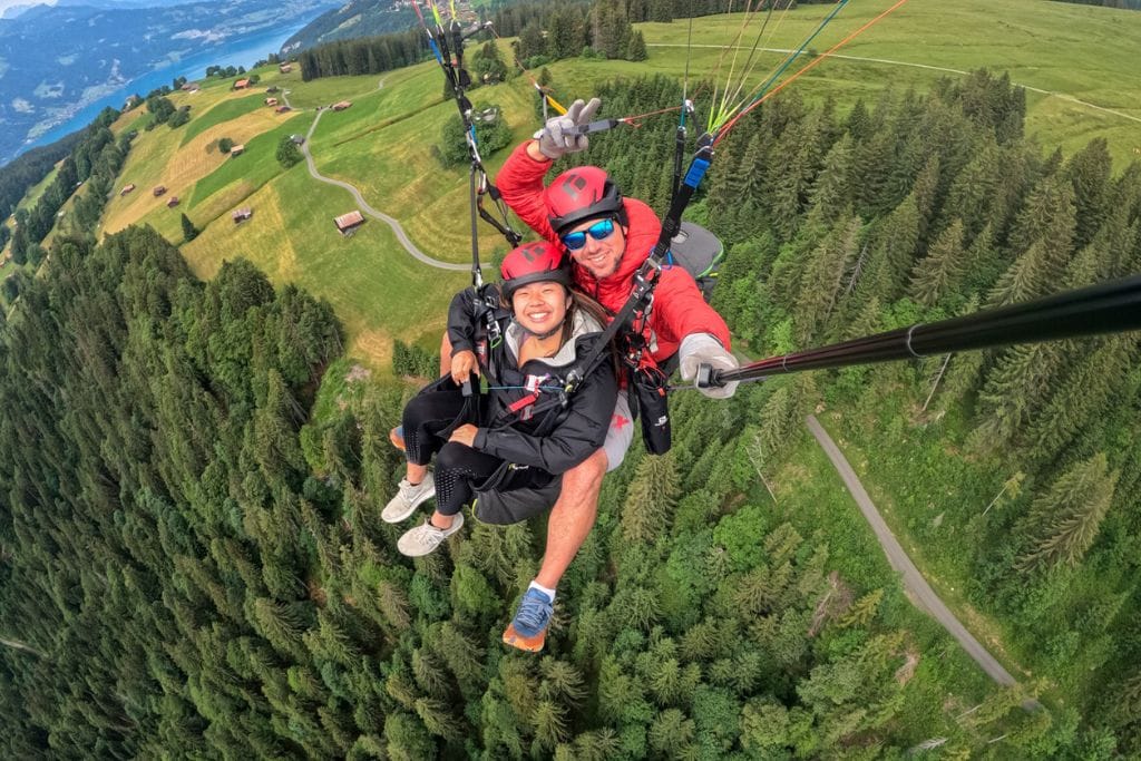 A picture of Kristin and her pilot smiling in the selfie while paragliding above Interlaken. You can see the verdant trees on the mountainside below.
