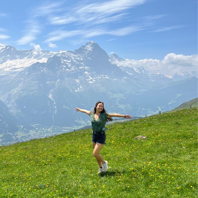 A picture of Kristin beaming with her arms out as she takes in the stunning views from the top of Grindelwald First in Switzerland!