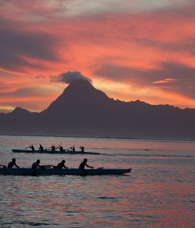 A picture of locals paddling in traditional outrigger canoes at sunset in Tahiti.
