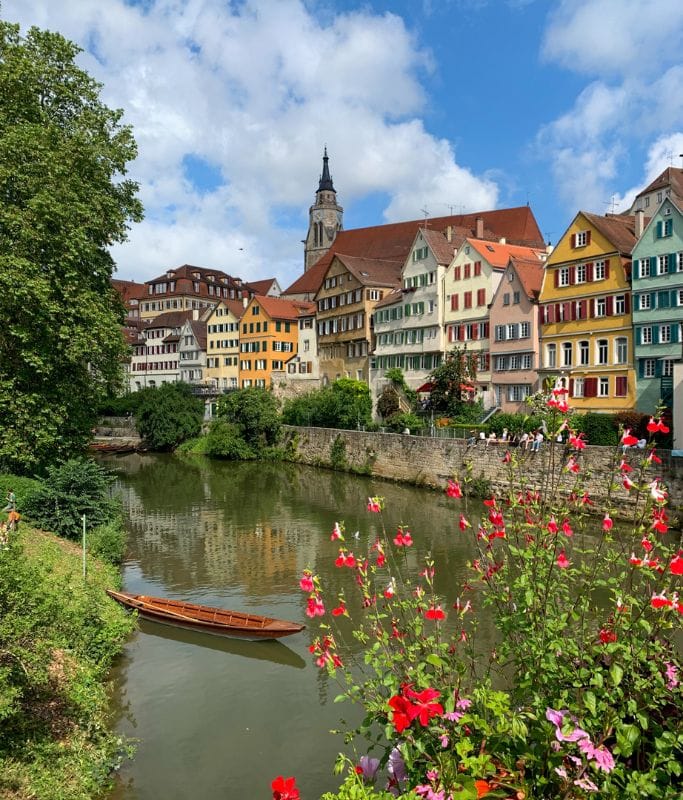 A picture of the iconic Neckarbrücke in Tubingen. You can see a Stocherkähne resting in the water, the colorful buidlings in the background, and all kinds of blossoming flowers in the front. Seeing this gorgeous view of Tubingen is a must things to do in the city.