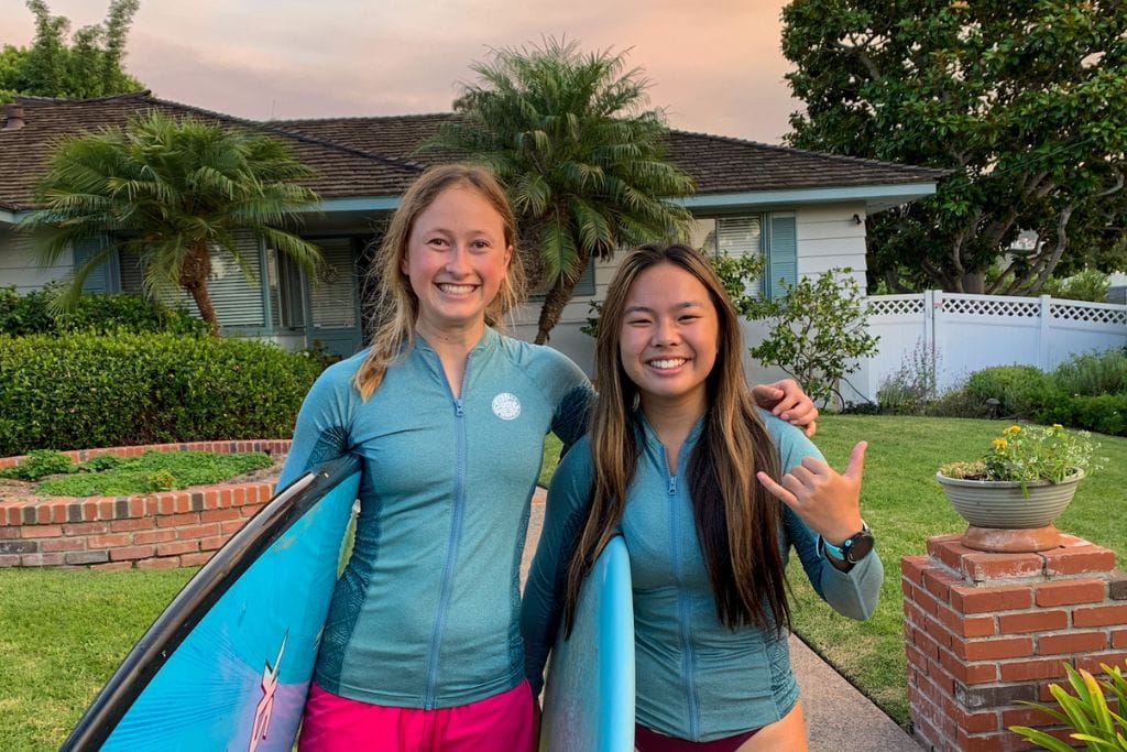 A picture of Kristin and her friend smiling with their surfboards as they get ready to surf.